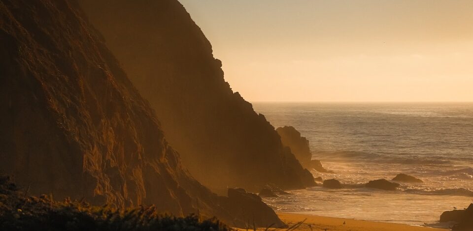 Golden hour illuminates a rugged coastline with ocean waves.
